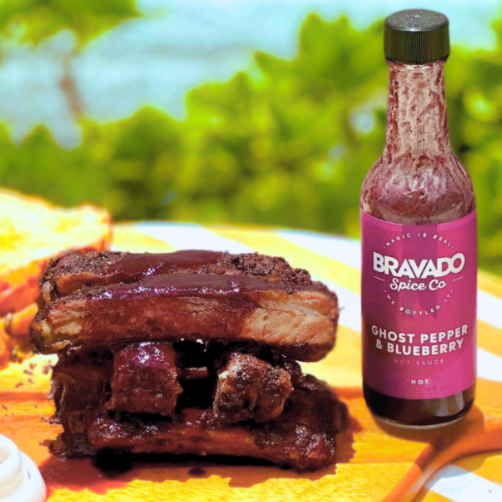 Glazed BBQ ribs stacked next to Bravado Spice Ghost Pepper & Blueberry hot sauce.