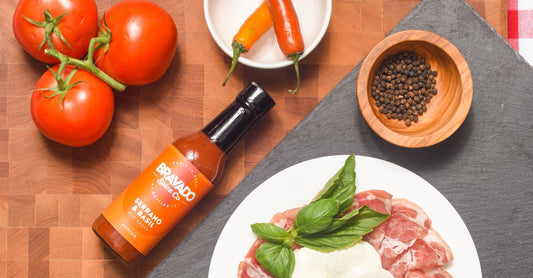 Bravado Spice’s Serrano & Basil Hot Sauce bottle on a wooden board with peppers, red chilis, tomatoes, and prosciutto.
