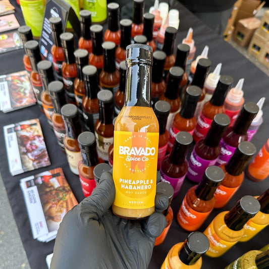 A gloved hand picks Bravado Spice Co.’s Pineapple & Habanero hot sauce at a display table with other hot sauces.